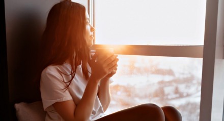 Retrato completo de uma jovem bonita vestindo camiseta branca e calças está sentado à luz do sol perto da janela com café e olhando para a cidade

