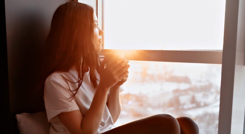 Retrato completo de uma jovem bonita vestindo camiseta branca e calças está sentado à luz do sol perto da janela com café e olhando para a cidade
