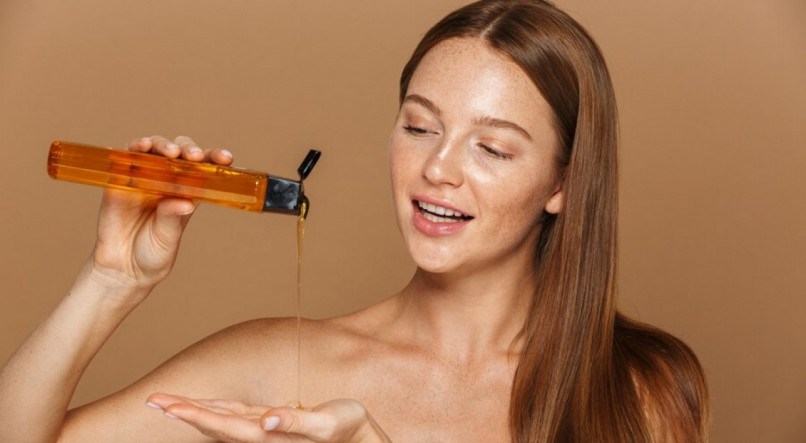 Retrato da beleza de uma jovem sorridente com longos cabelos vermelhos mostrando o frasco de shampoo isolado sobre uma parede bege