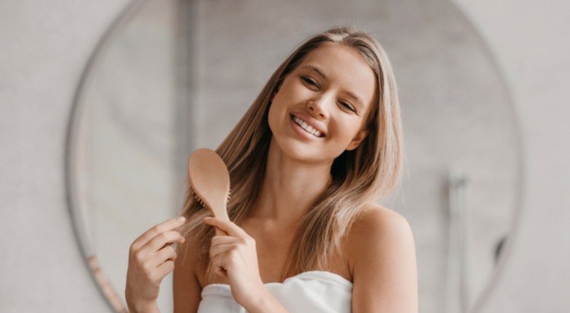 Haircare concept. Pretty lady brushing her beautiful hair with comb while looking at her reflection and smiling in mirror in bathroom interior