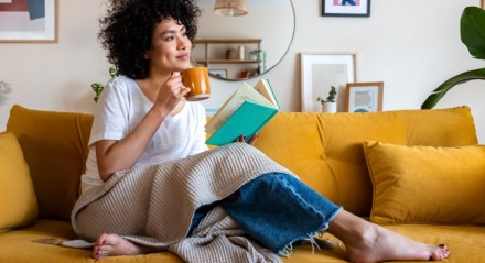 Mulher lendo um livro e tomando um café sentada em um sofá amarelo da sala