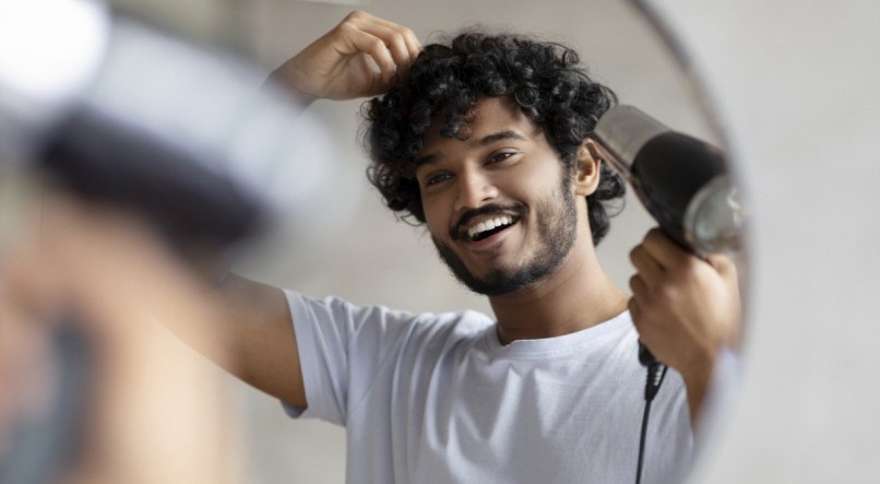 Excited indian man using hairdryer after shower, drying his curly hair, making hairdo, taking care of himself in the morning, smiling at his reflection in mirror. Everyday hygiene concept