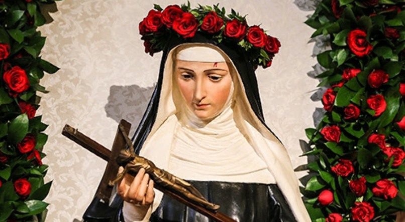 Imagem ilustra Santa Rita de Cássia no centro segurando uma cruz, com uma coroa de rosas vermelhas na cabeça, e outras rosas espalhadas pelos lados
