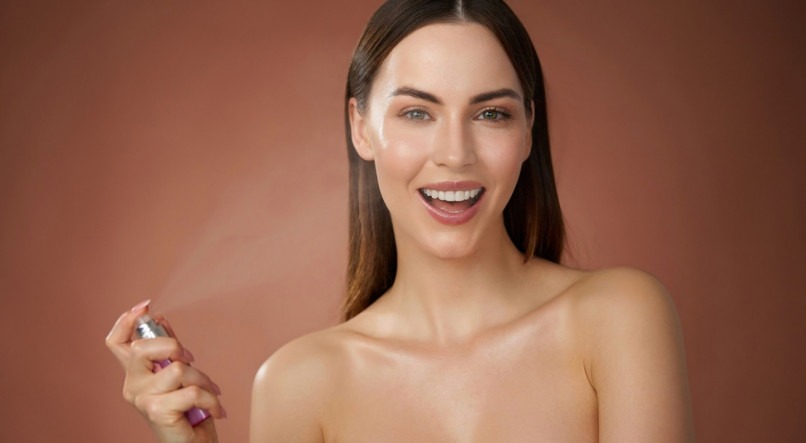 Positive young female with bare shoulders smiling and looking at camera while spraying refreshing water standing on brown background in studio