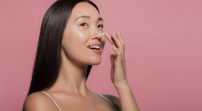 Close up of a youthful female model applying moisturizer to her face. Young korean woman putting moisturizer cream on her pretty face against pink background.