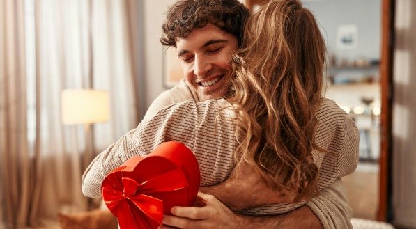 Happy Valentine's Day. A man gives a heart-shaped gift box to his beloved woman in the living room at home, the woman hugging him tenderly. Romantic evening together.