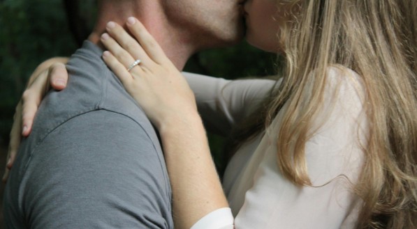 Homem de camisa cinza beijando mulher de camisa branca e aliança no dedo. Imagem ilustrativa
