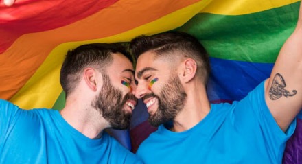Imagem ilustra casal gay com a bandeira LGBT, em alusão ao Dia Internacional do Orgulho