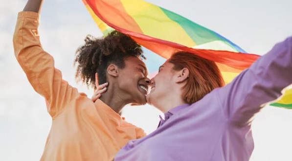 Imagem ilustra casal de mulheres se beijando com a bandeira do arco-íris, o símbolo do orgulho LGBT