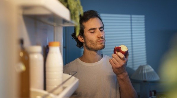 Imagem ilustrativa de um homem comendo em frente à geladeira.