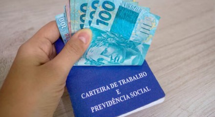 Imagem ilustra uma mão segurando dinheiro sobre a carteira de trabalho em alusão ao PIS/Pasep, o benefício do trabalhador