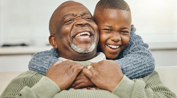 Imagem ilustra avô negro sorrindo e o neto o abraçando por trás