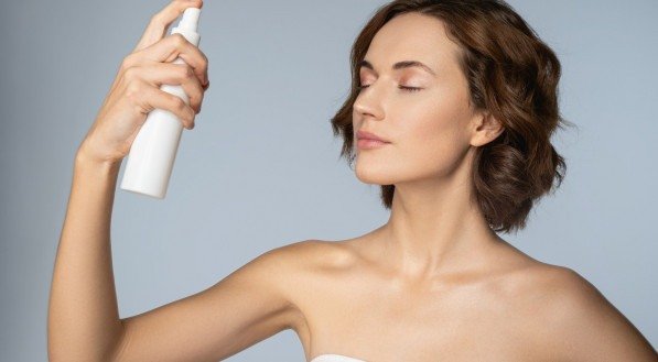 Young beautiful woman is spraying cosmetic treatment on skin for moisturising isolated on grey background