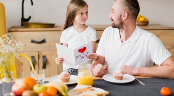 Imagem ilustrativa de café da manhã especial de Dia dos Pais, com filha pequena lendo cartão comemorativo para o pai 