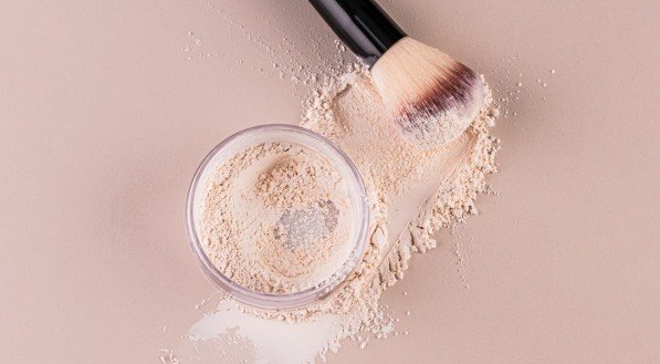 Glass jar with loose powder, swatch powder and makeup brush, applying powder on face on beige background. Concept of cosmetics , makeup. Top view