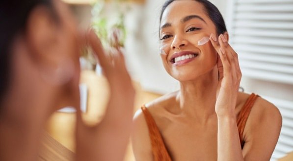 Portrait of smiling latin woman applying cream on her face while looking in mirror in her bathroom. Young multiethnic woman applying moisturizer on her cheek while looking in the mirror. Happy mexican girl using lotion every morning.