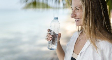 Imagem ilustrativa de uma mulher tomando água na praia para evitar a desidratação