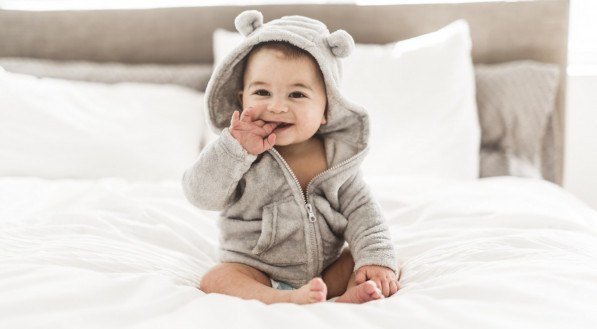 Foto de um bebê sentado na cama vestindo um roupão cinza com orelhas temáticas.