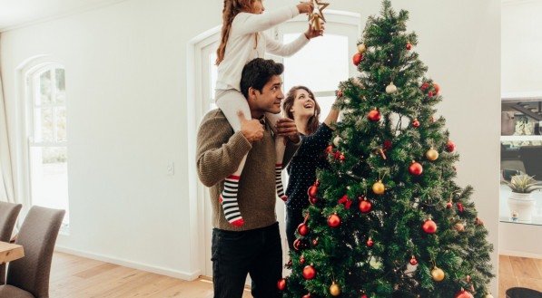 Imagem de uma família decorando uma árvore de Natal. Jovem com sua filha nos ombros ajudando-a a decorar a árvore de Natal