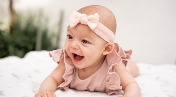 Retrato em close-up de uma bebê rindo com a boca aberta vestida com uma camisa babada e um laço rosa na cabeça deitada em um cobertor.