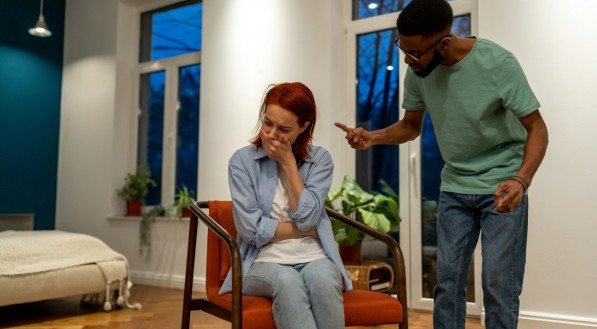Imagem de um momento tenso em um ambiente doméstico, onde um homem está discutindo e gesticulando para uma mulher sentada.