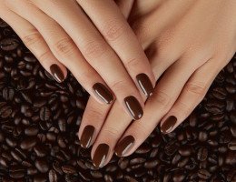 Unhas pintadas de tom marrom, com o fundo de café atrás das mãos.