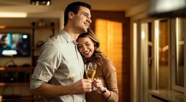 Imagem de um casal feliz abraçado e brindando com taças de espumante em casa