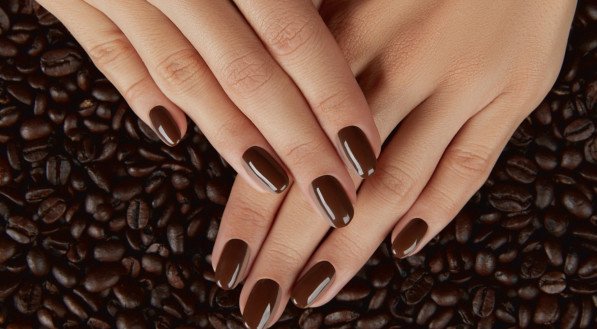 Unhas pintadas de tom marrom, com o fundo de café atrás das mãos.