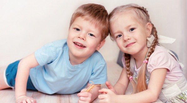 Irmãos gêmeos, um menino e uma menina, sorrindo e posando para foto em fundo claro.