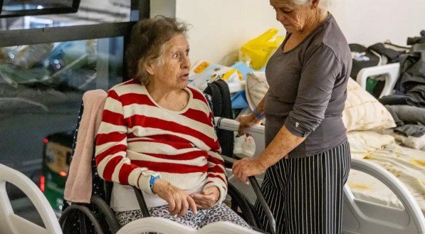 Imagem de uma mulher idosa sentada em uma cadeira de rodas, acompanhada por outra idosa em pé, que segura apoio da cadeira