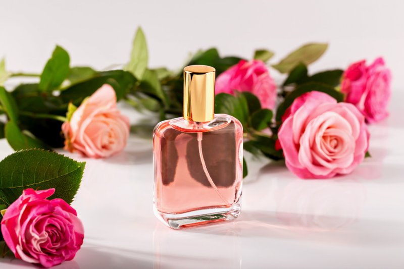 Frasco de perfume de vidro com tampa dourada preenchida com líquido rosa, cercado por rosas cor-de-rosa frescas na superfície branca.