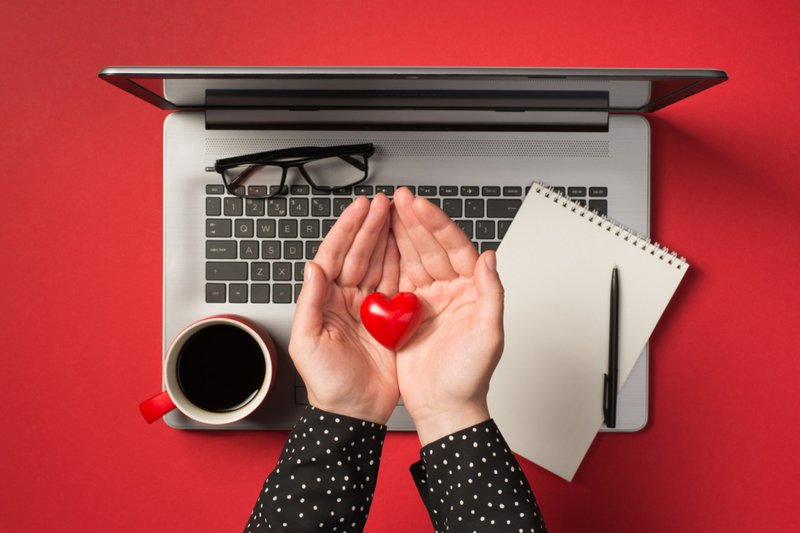 Imagem aérea de copos, caneta, bloco de notas, laptop cinza, copo de café e mãos segurando um coração vermelho isolado no cenário vermelho