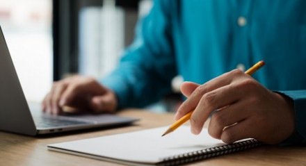 Um homem escreve em um caderno com um lápis. Ele está concentrado no trabalho e com um humor sério. O caderno está sobre uma mesa de madeira, e há um laptop por perto.