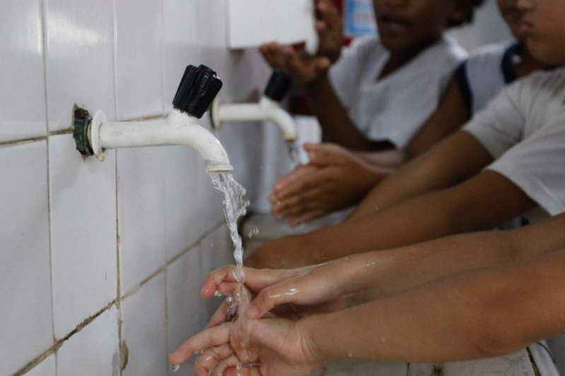 Imagem de crianças lavando as mãos em pia coletiva, em ambiente escolar