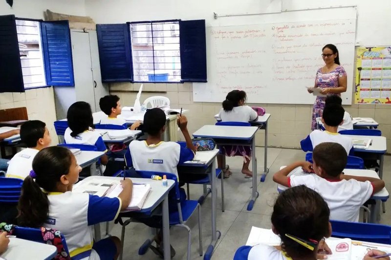 Imagem de uma sala de aula com alunos sentados em suas carteiras, enquanto a professora está em pé à frente da turma, conduzindo a aula