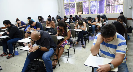 Pessoas sentadas em cadeiras de salas de aula realizando prova