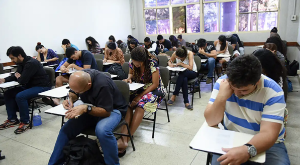 Pessoas sentadas em cadeiras de salas de aula realizando prova. Imagem ilustrativa para falar do Encceja