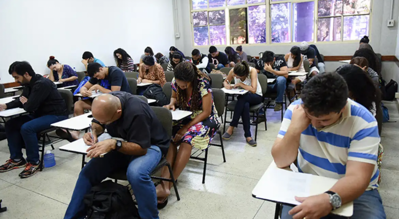 Pessoas sentadas em cadeiras de salas de aula realizando prova. Imagem ilustrativa para falar do Encceja