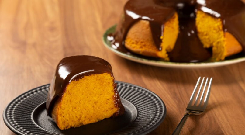 fatia de bolo de cenoura com cobertura de chocolate em primeiro plano, está o bolo inteiro.