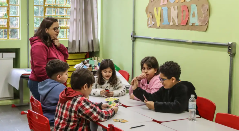 Crianças em escola jogam jogo de cartas em mesa, professora observa em pé. Imagem ilustrativa para falar sobre escolas no Rio Grande do Sul