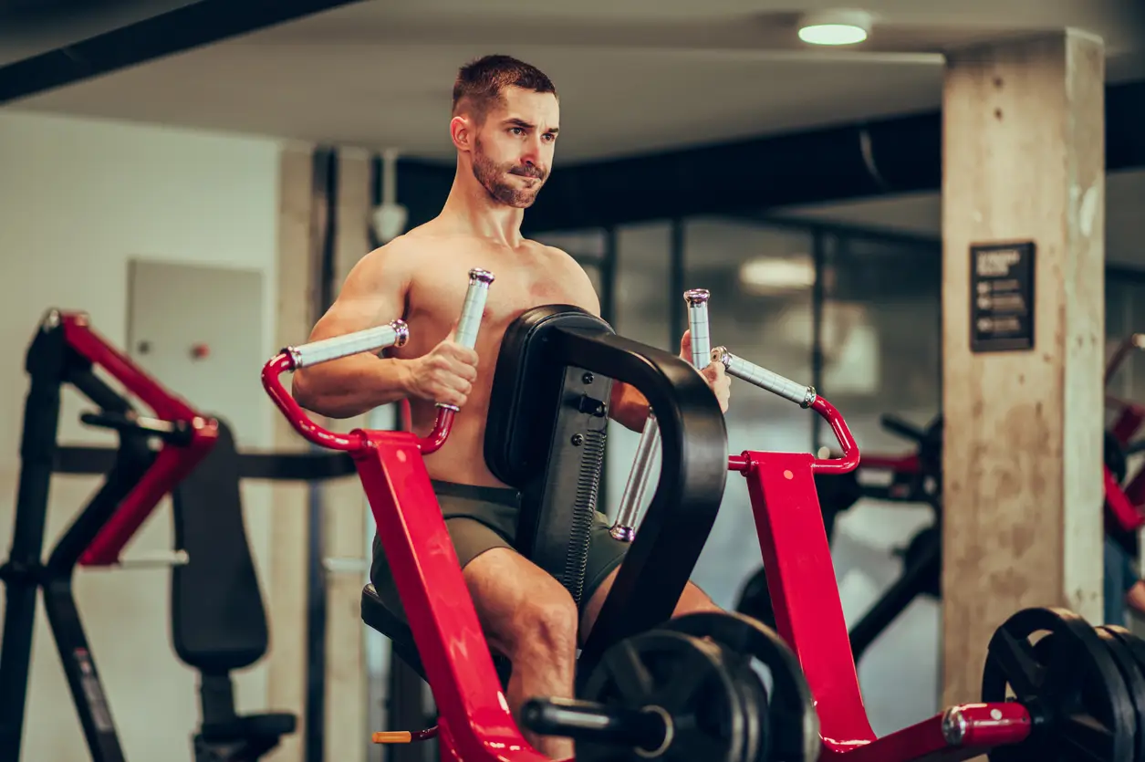 Imagem ilustrativa de um jovem esportista musculoso e sem camisa está se exercitando em uma máquina de Remada Máquina Aberta na academia