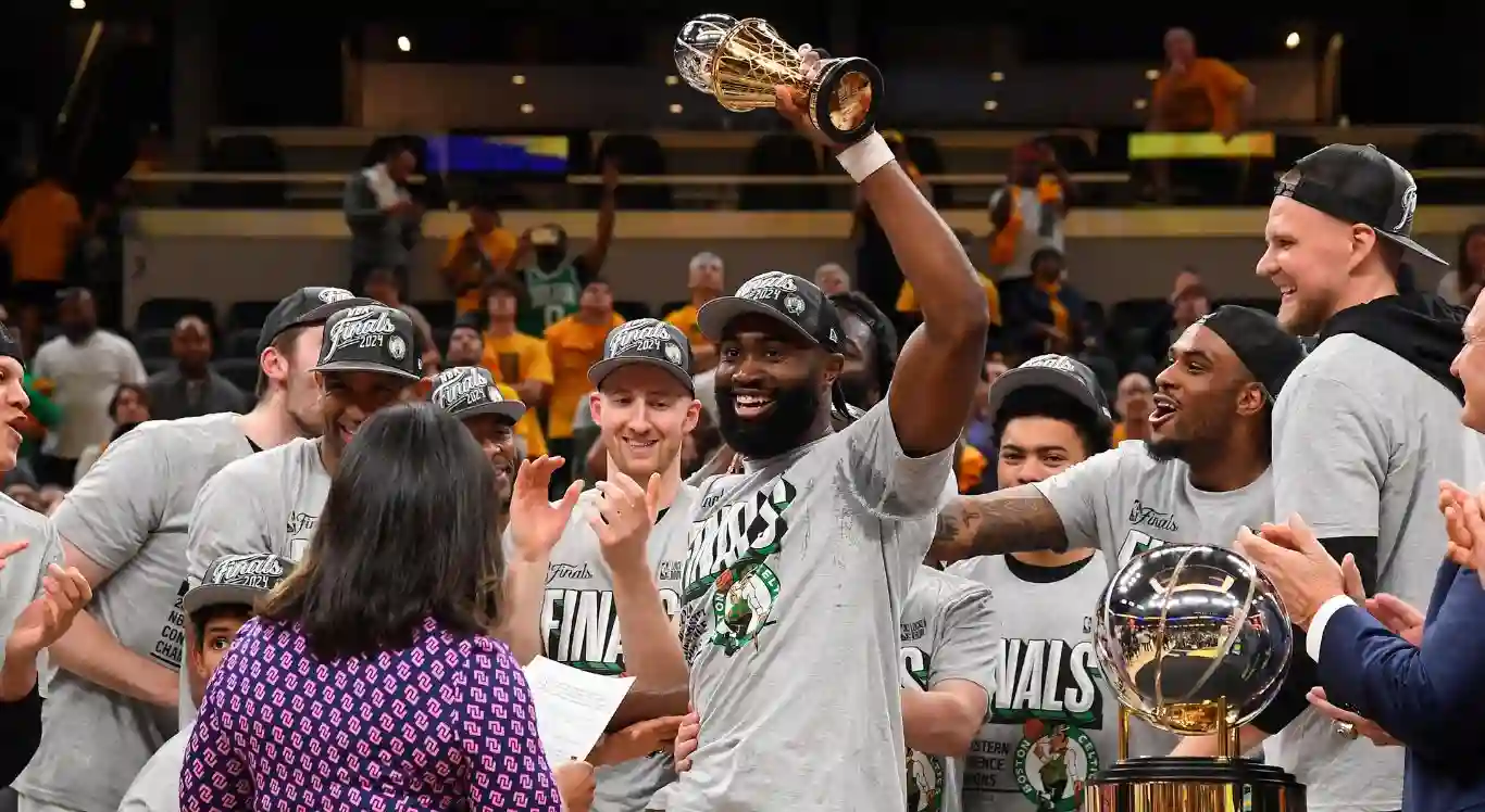 Imagem de Jaylen Brown com trof&eacute;u de MVP das Finais do Leste e time do Boston Celtics celebrando vit&oacute;ria na NBA