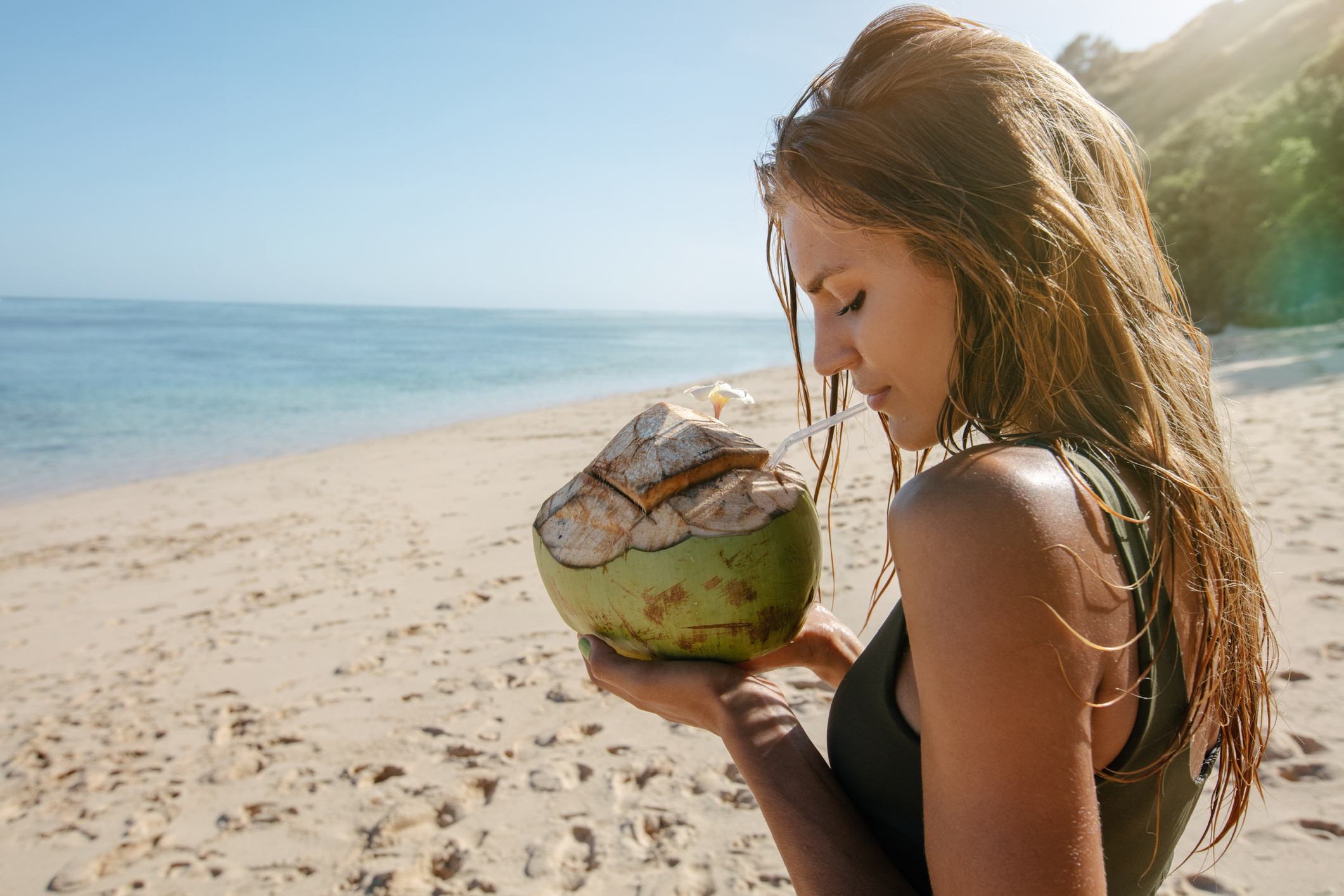 Mulher tomando água de coco na praia com cabelos molhados.