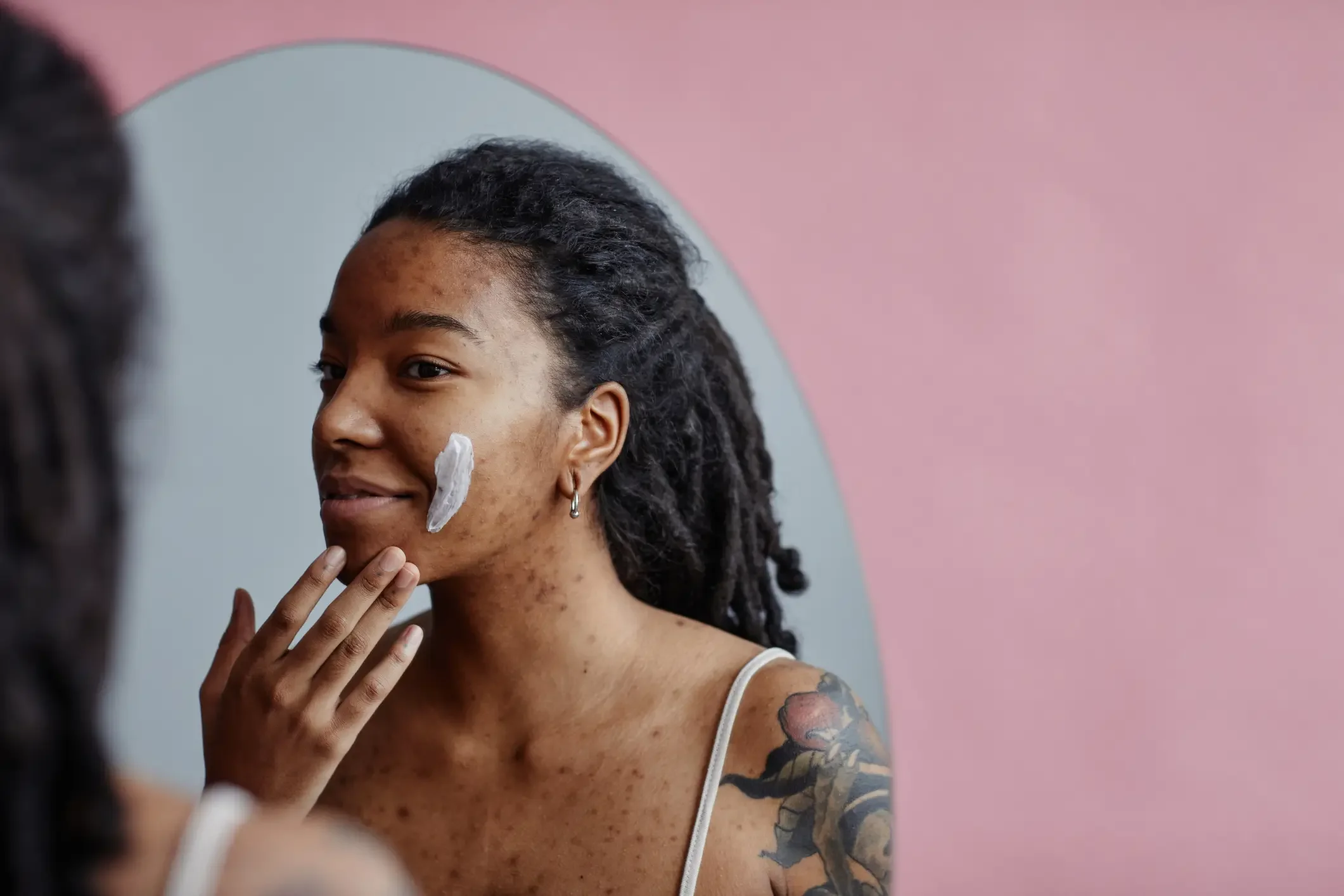 Imagem ilustrativa de mulher negra nova com cicatrizes de acne usando creme facial olhando no espelho.