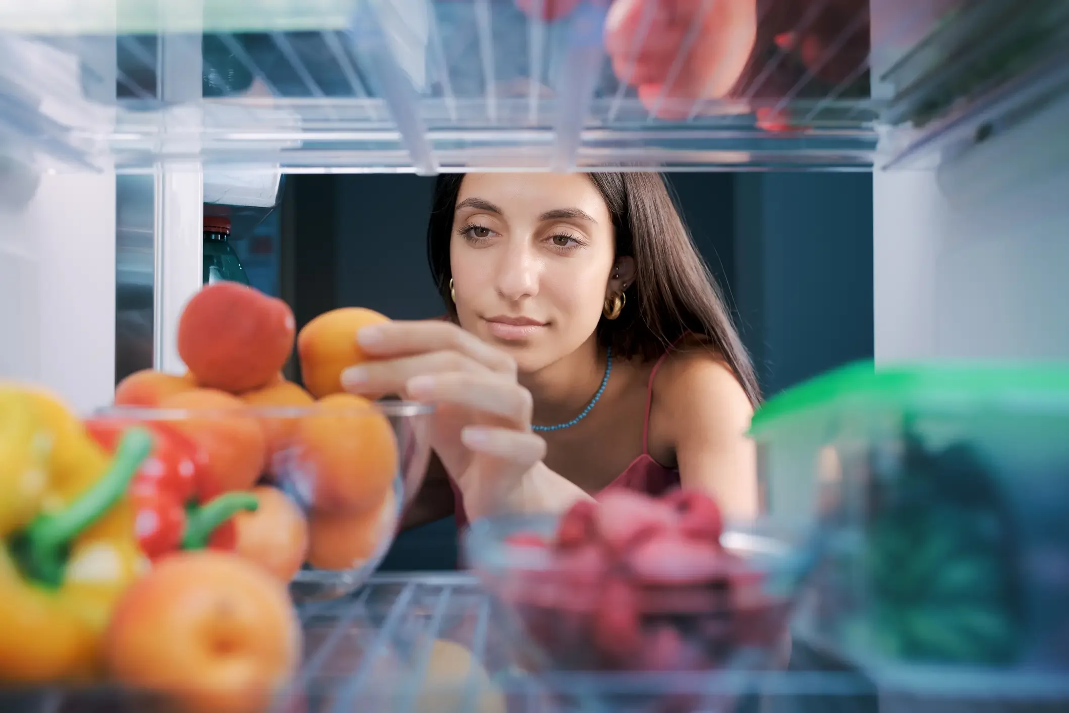 Imagem de uma mulher olhando frutas na geladeira de noite.