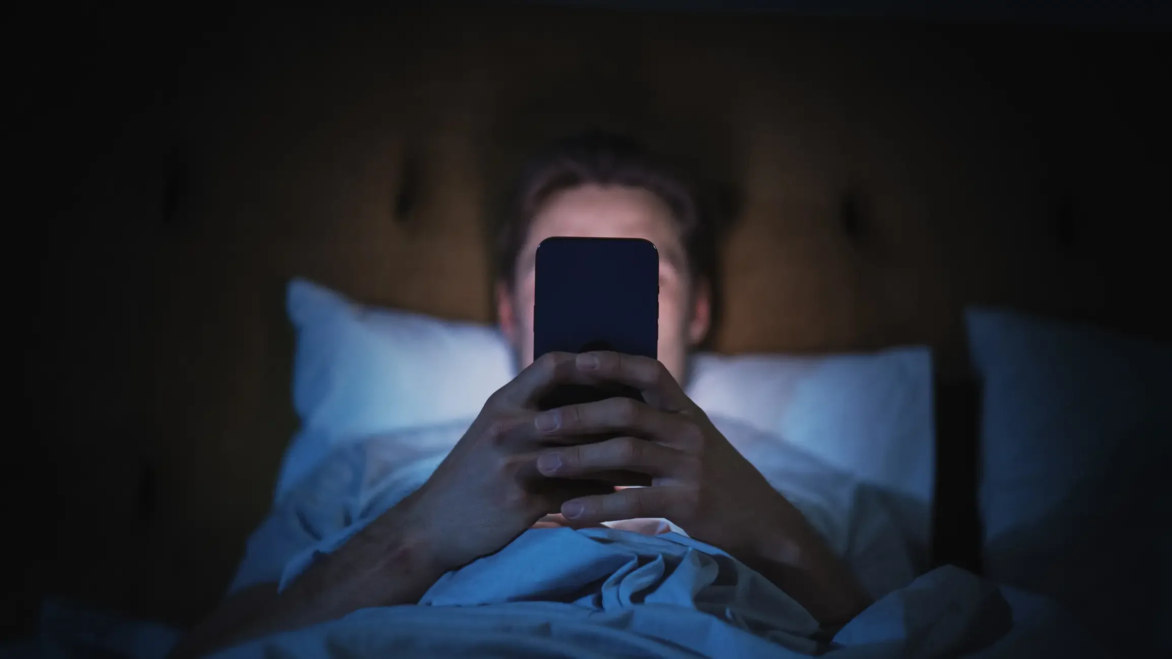 Imagem de homem com o celular ligado na cama de noite.