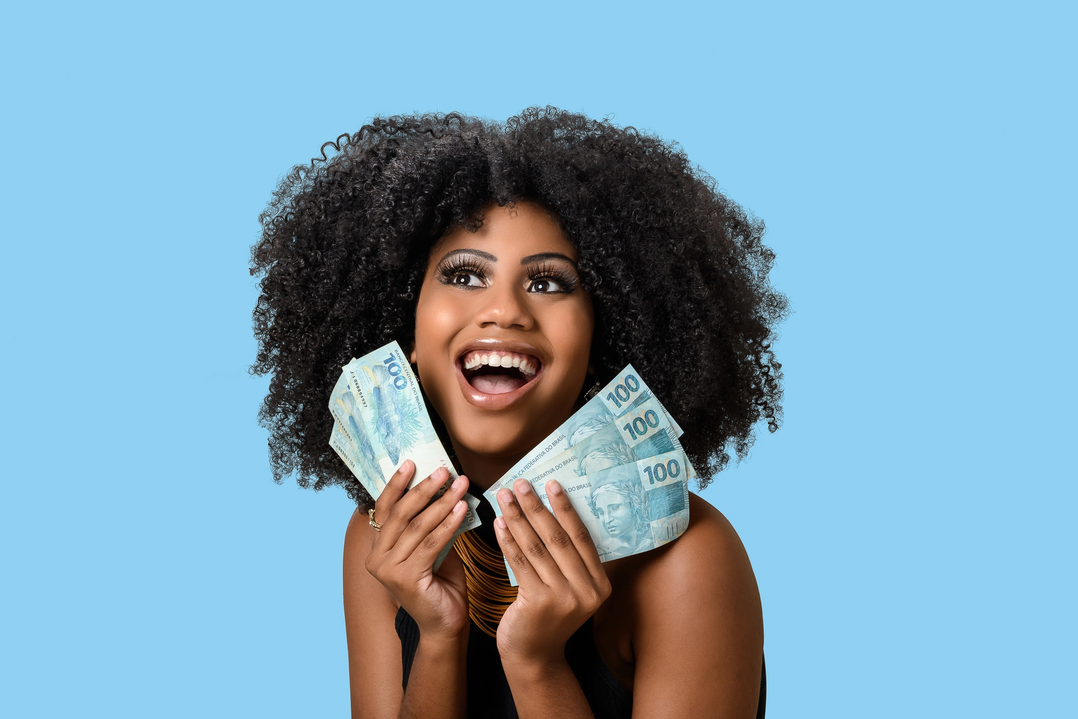 Jovem negra sorrindo segurando contas de dinheiro brasileiro, positivamente surpreendida, isolada no fundo azul.
