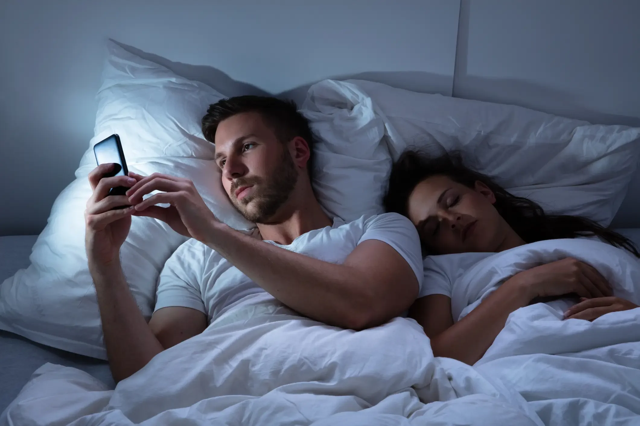 Imagem ilustrativa de homem usando telefone celular enquanto sua esposa sentada na cama