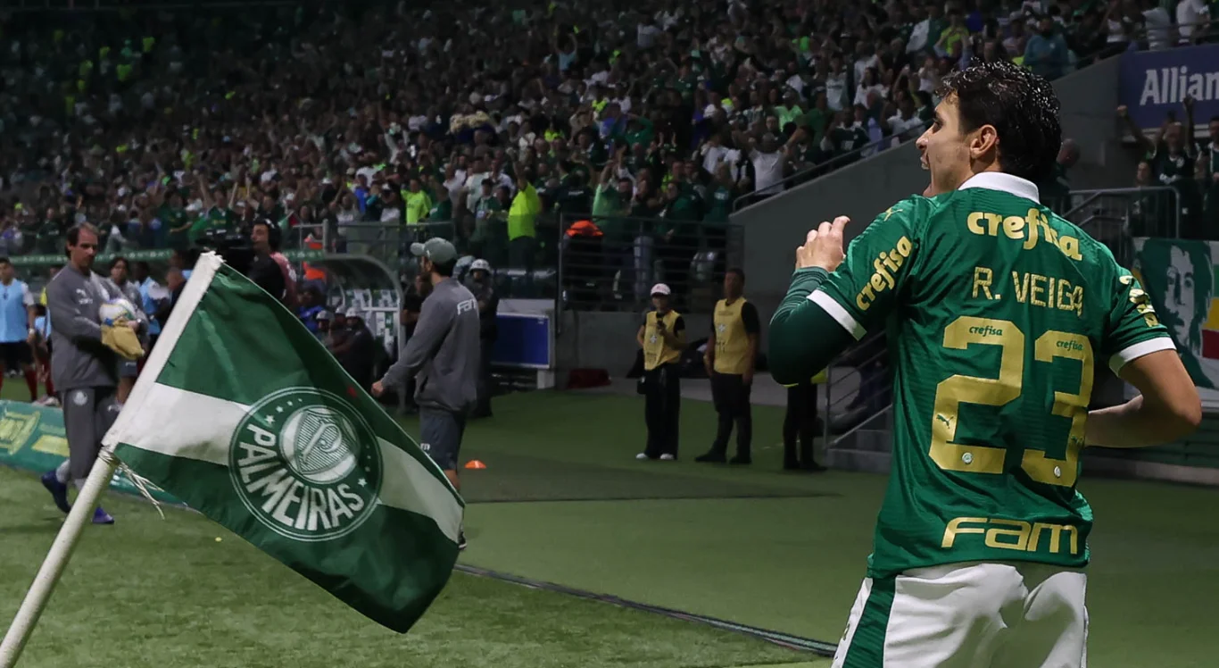 Imagem de Raphael Veiga com a torcida ao fundo e a bandeira do Palmeiras