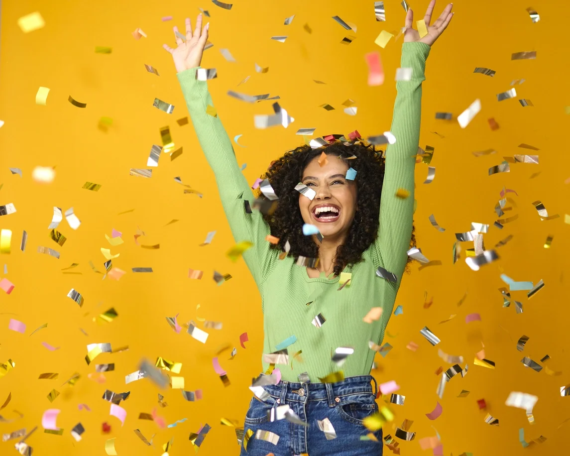 Foto de estúdio de mulher animada comemorando grande vitória banhada em confetes de tinsel no fundo amarelo
